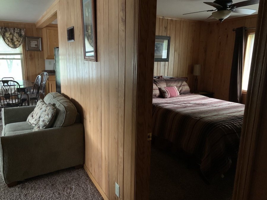 Inside a cabin: knotty pine walls, a small kitchen and dining table, and a bedroom with a queen bed through the doorway