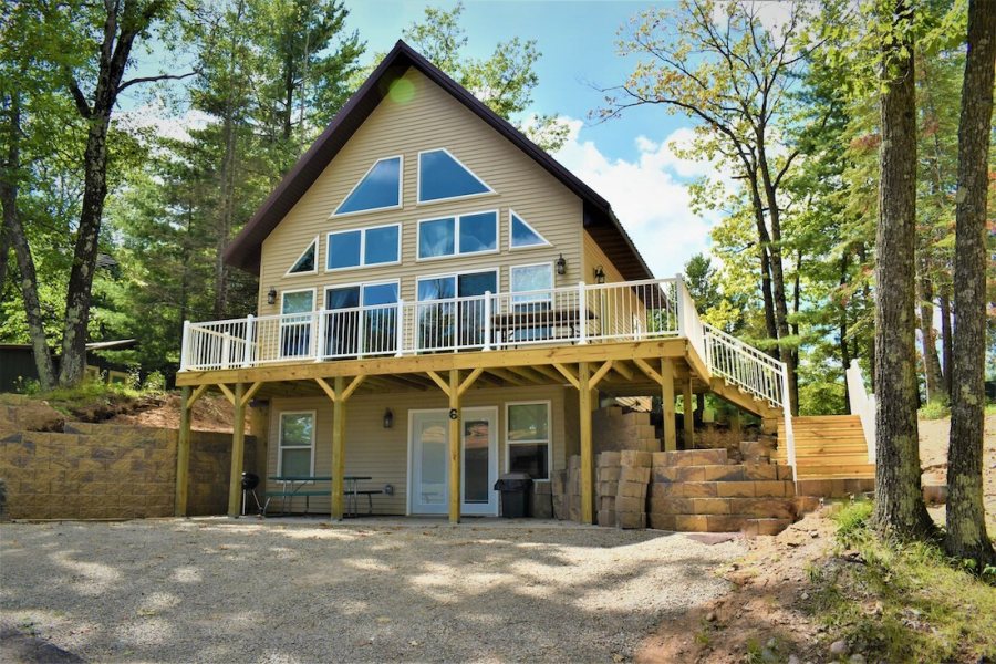 A cabin with a steep A-frame window wall and a raised wrap-around deck, set among birch and pine above a gravel drive