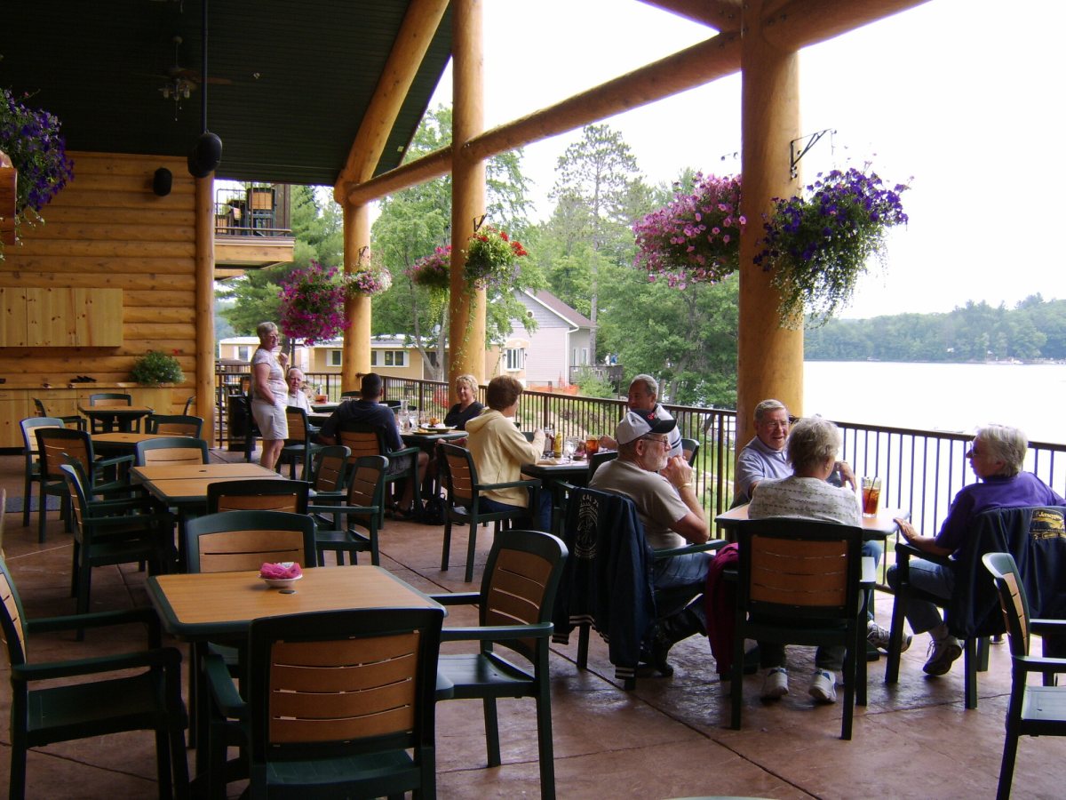 Guests eating at tables on the covered lakeside deck, log columns and hanging baskets framing the water
